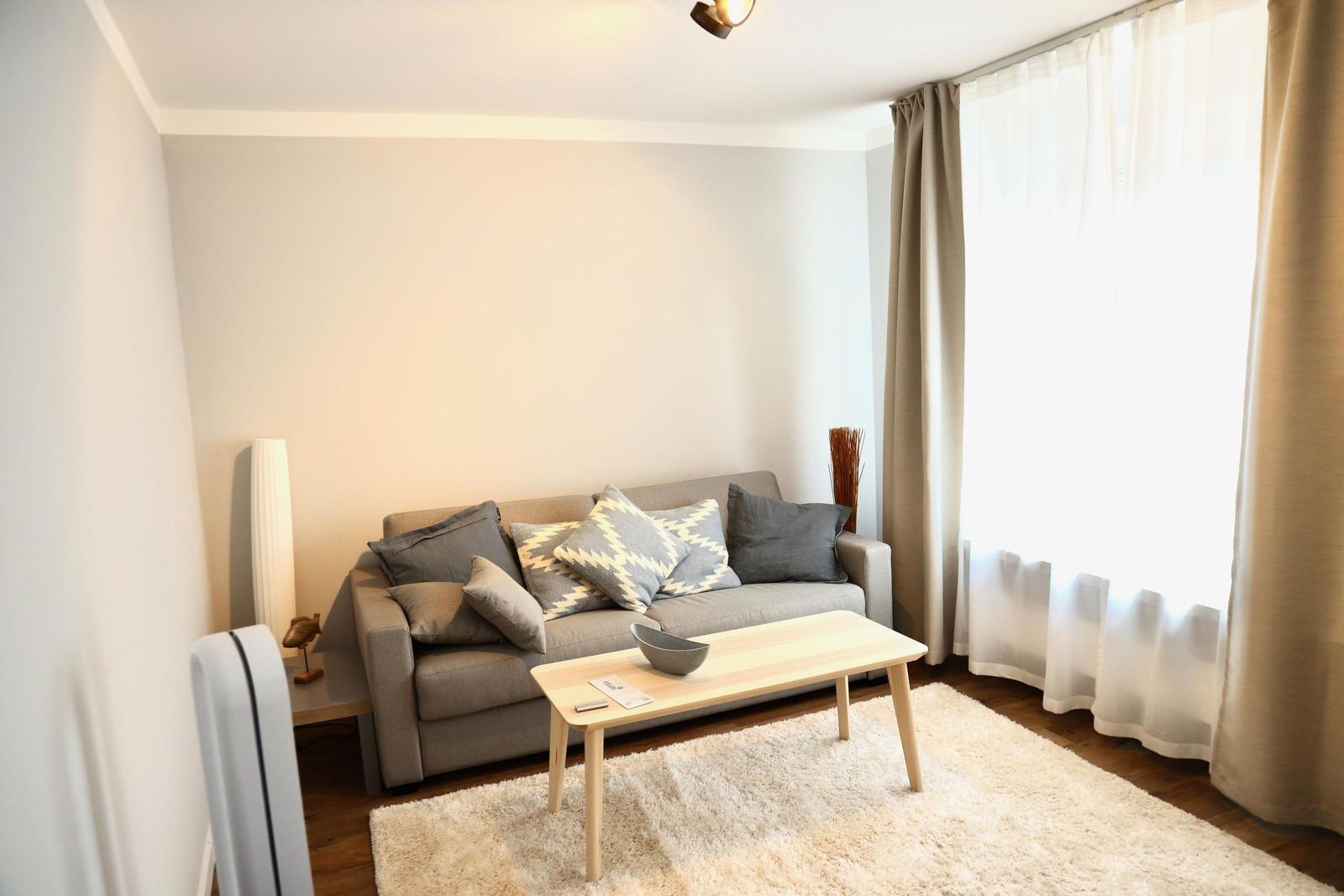 Ein Wohnzimmer mit grauem Sofa, Holztisch und Fenster mit Vorhängen.