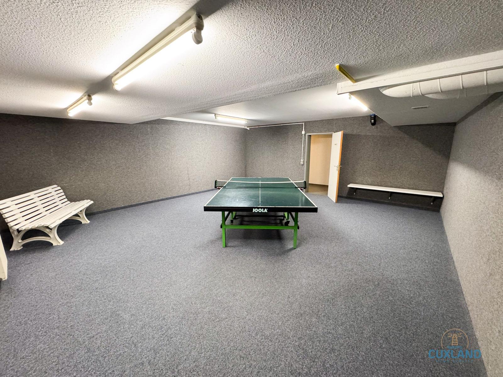 Raum mit Tischtennisplatte, Sitzbänken und Deckenleuchten.