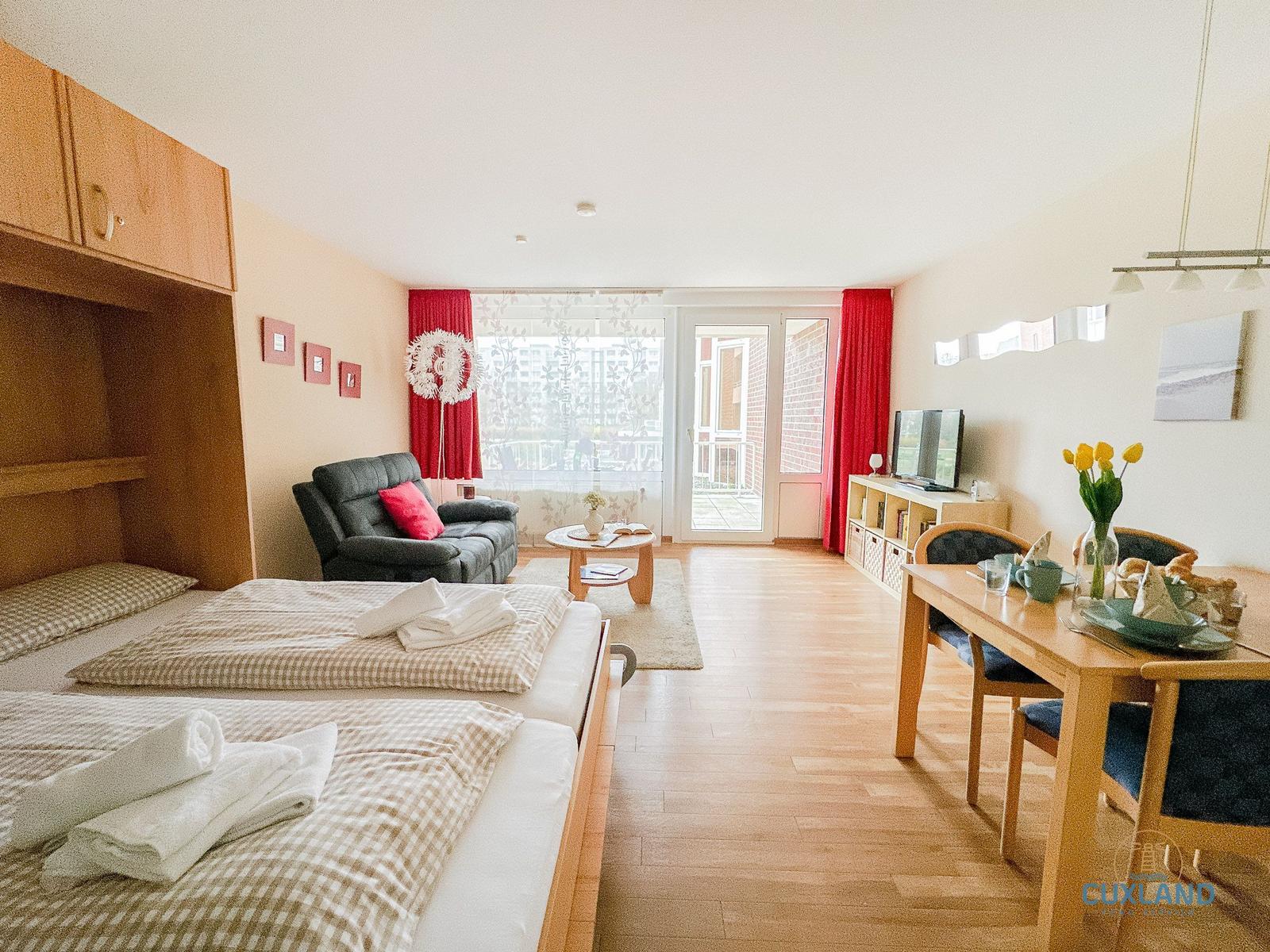 Zwei Betten, Sofa, Esstisch und Balkon mit roten Vorhängen.