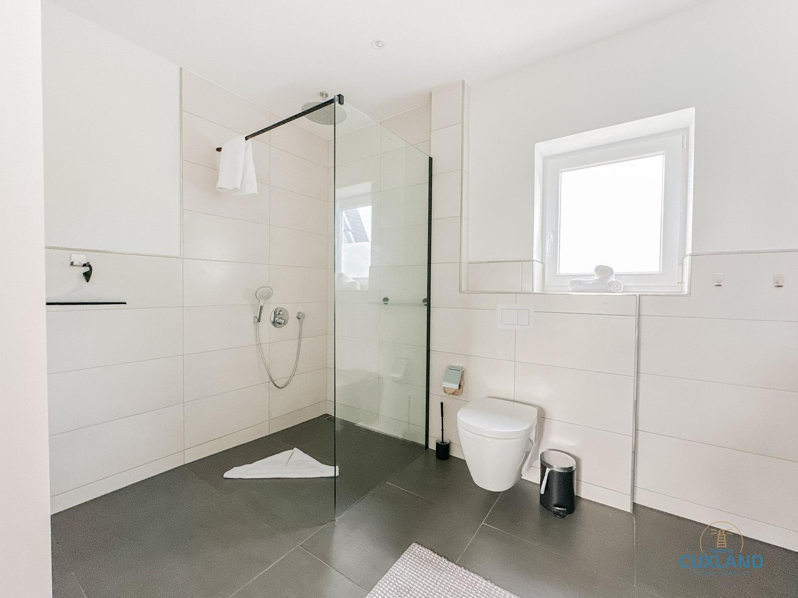 Badezimmer mit Dusche, Toilette und Fenster. Fliesenboden und weiße Wände.