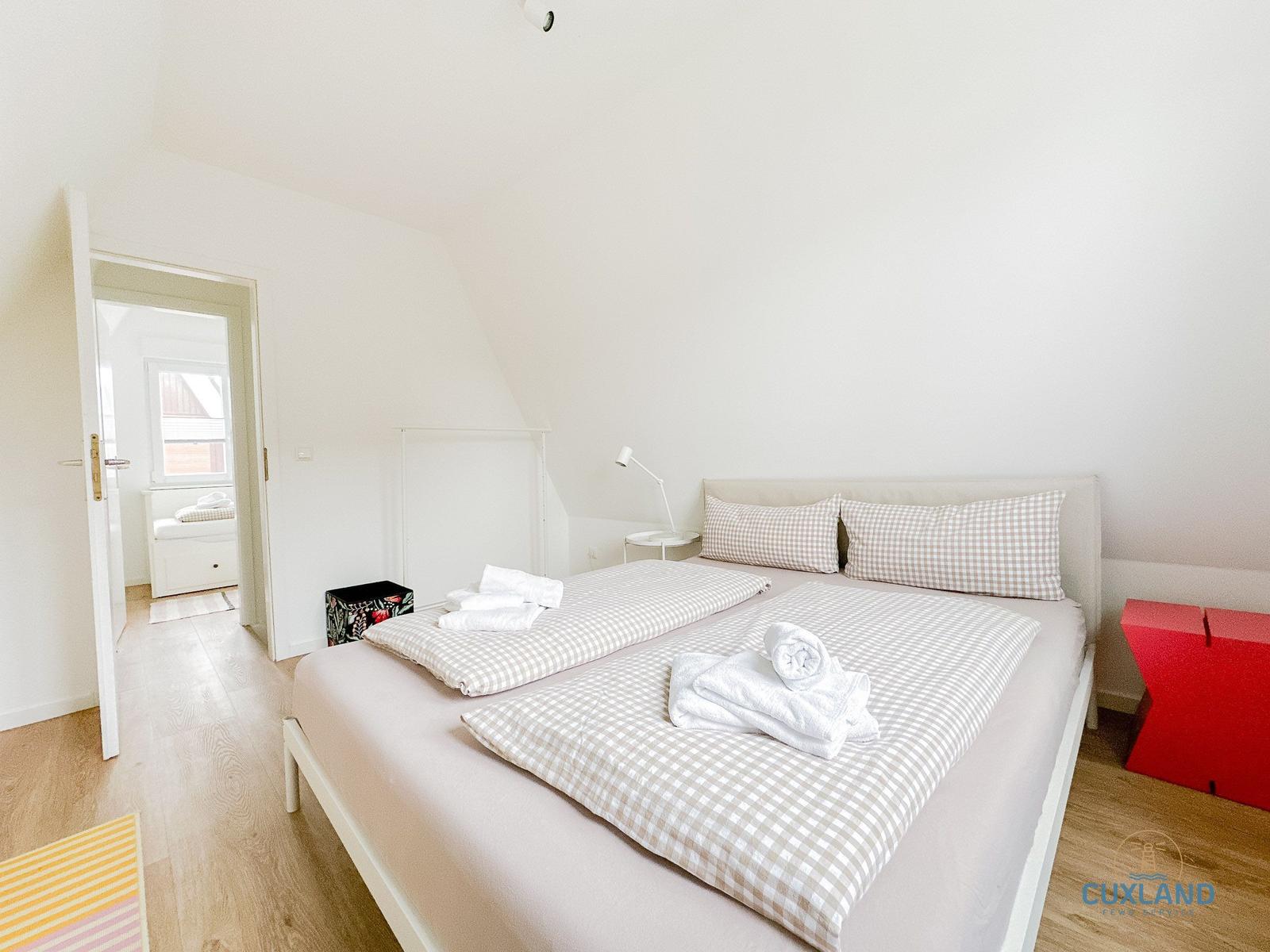 Schlafzimmer mit Doppelbett, weißen Wänden und Holzboden. Durchgang zu einem Fensterbereich.