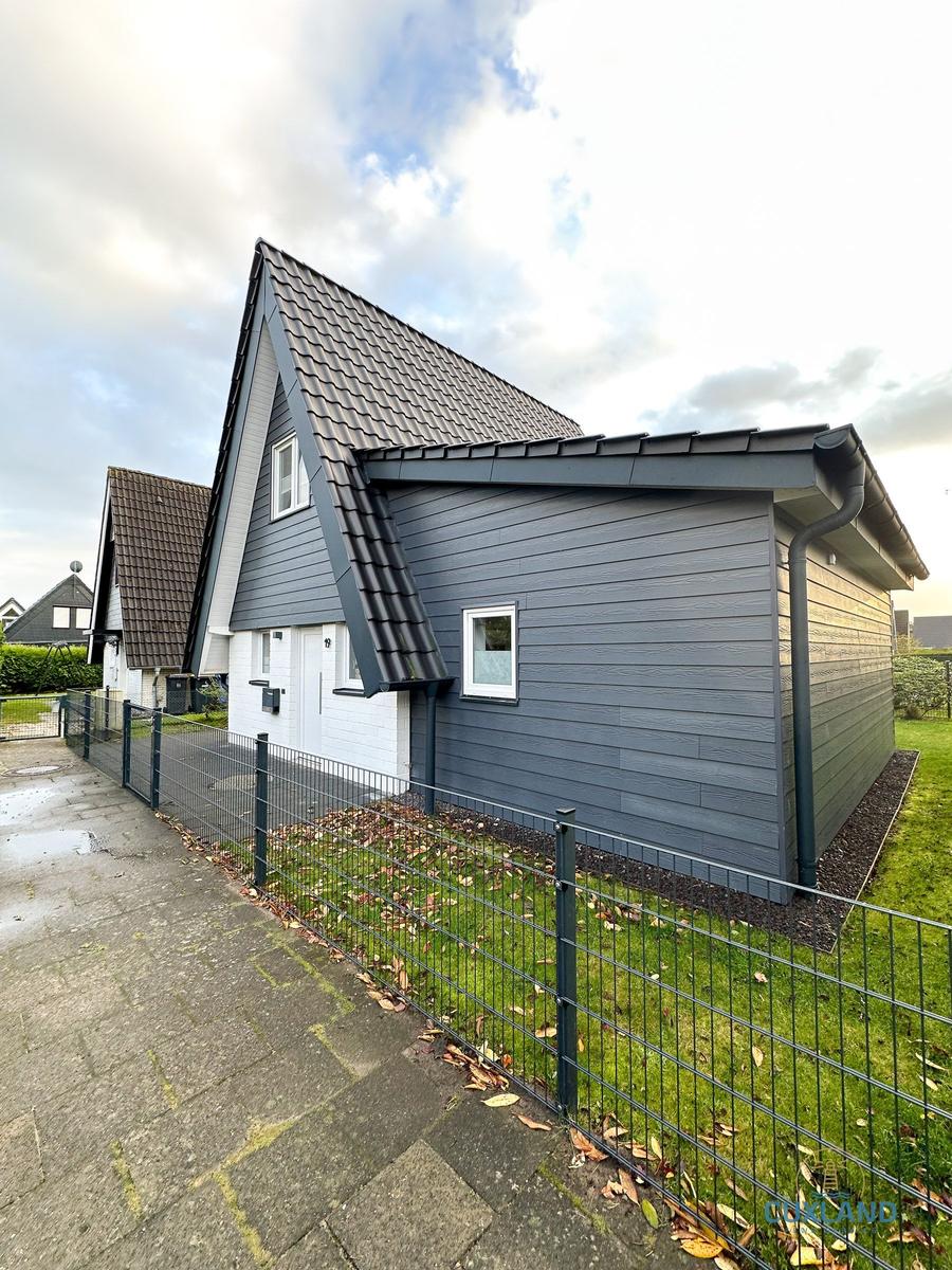 Moderne Villa mit grauer Fassade und weißer Tür. Grünfläche und Zaun sind sichtbar.