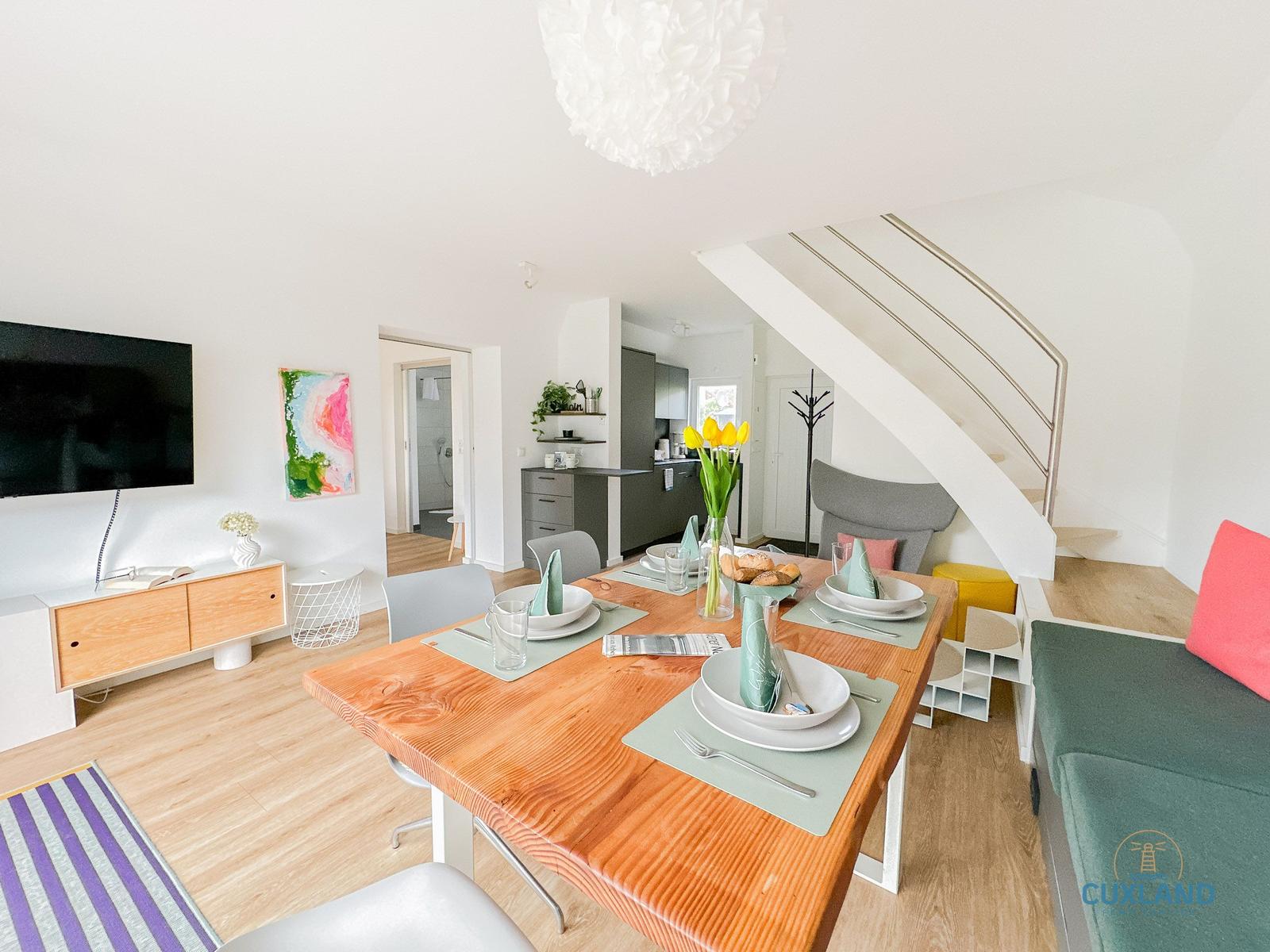 Essbereich mit Holztisch, Stühlen und Blumen. Küche, Wohnbereich und Treppe sind sichtbar.