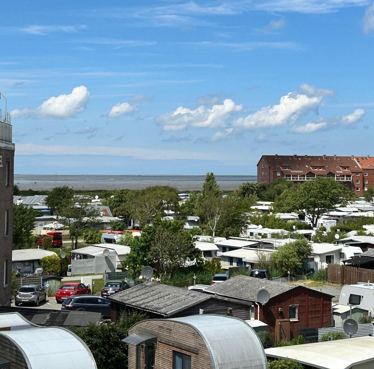 Panorama mit Campingplatz, Bäumen und Meer unter blauem Himmel mit weißen Wolken.