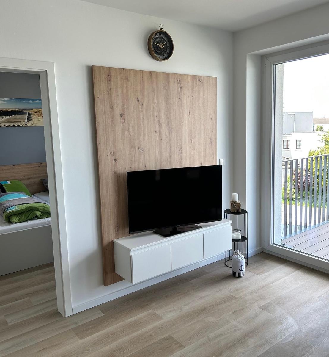 Wohnzimmer mit TV, Holzwand und Balkonzugang. Nebenraum mit Bett sichtbar.