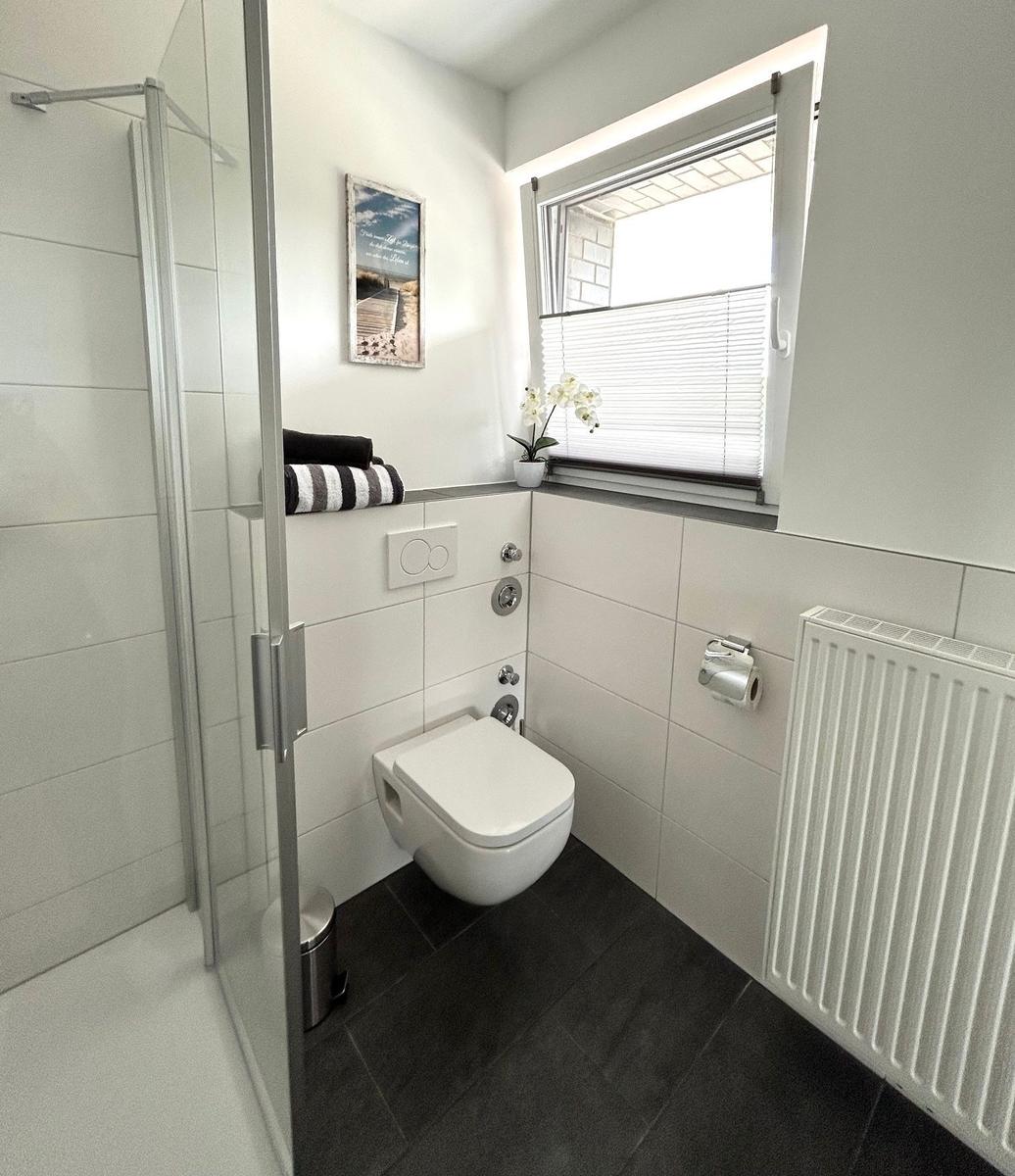 Badezimmer mit Dusche, Toilette und Fenster mit Jalousie.
