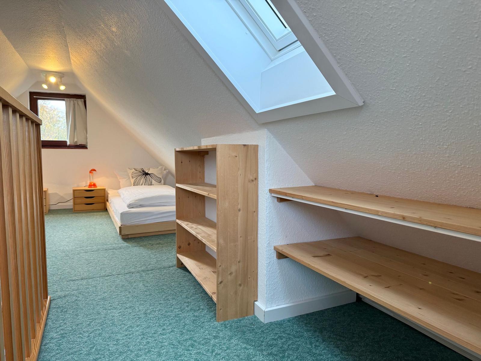 Bedroom with sloped ceilings, wooden shelves, and skylight. Bed, nightstand, and window are visible.