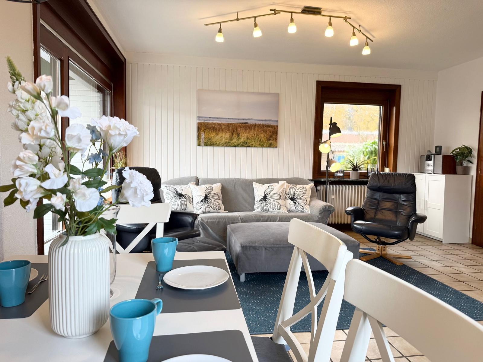 Living and dining area with sofa, chairs, and table. Windows and wall art visible.
