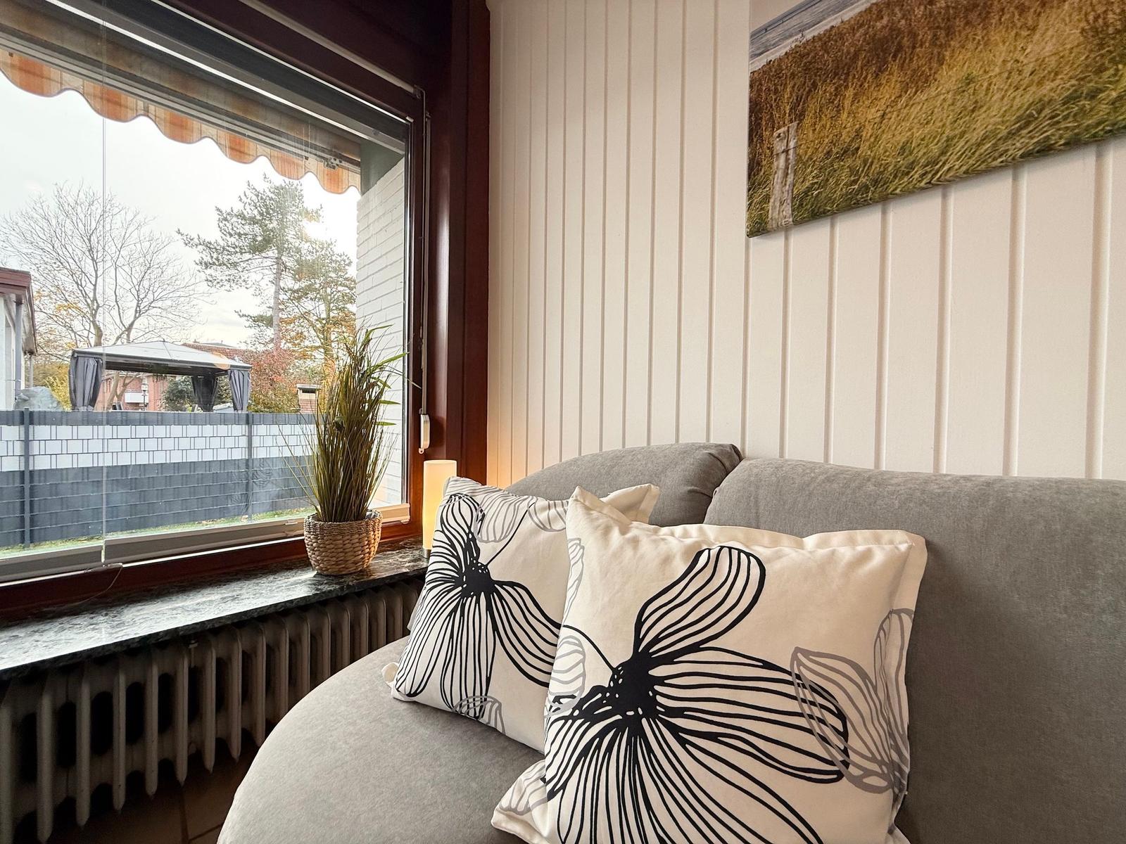 A gray seating area with cushions by a window overlooking a garden.
