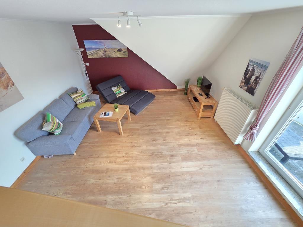 Wohnzimmer mit Sofa, Sitzbank, Couchtisch und Fernseher. Holzdielenboden und große Fenster.