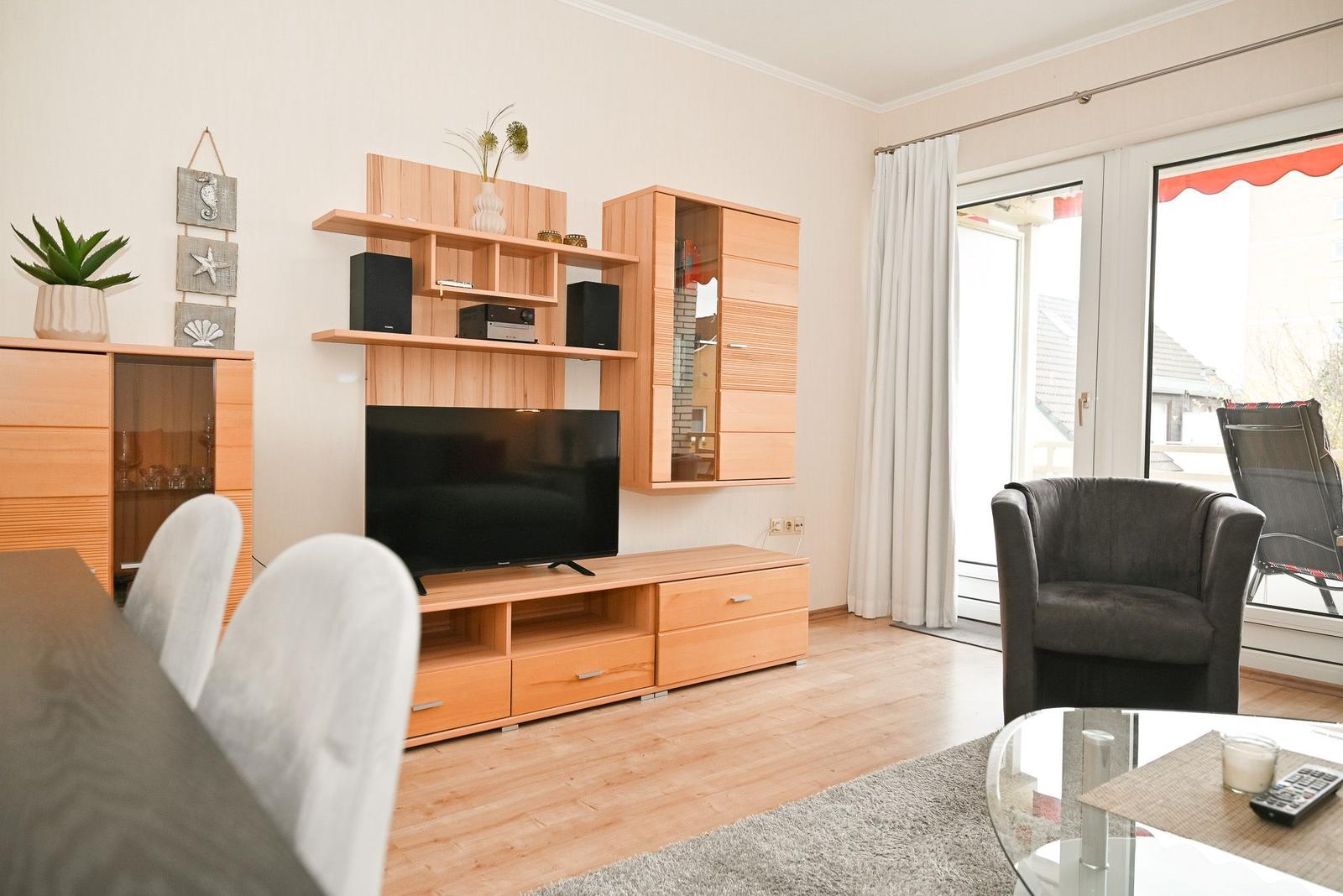 Wohnzimmer mit Holzmöbeln, Fernseher und Sitzgelegenheiten. Durchgang zur Terrasse mit Sicht auf die Umgebung.