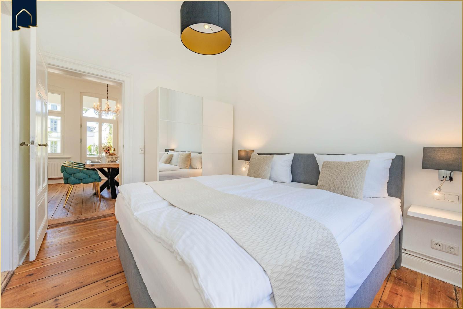 Schlafzimmer mit Doppelbett, Spiegelschrank und Holzboden. Durchgang zu Essbereich mit Tisch und Stuhl.