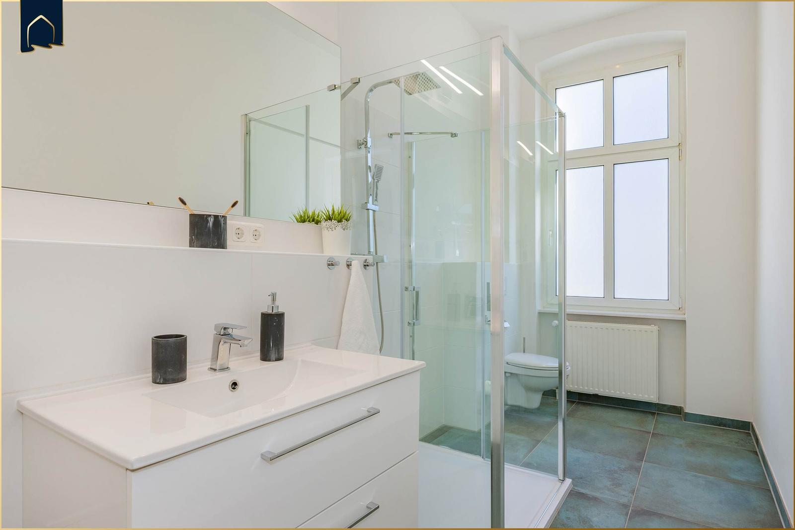 Badezimmer mit Dusche, Waschbecken und Toilette. Fenster sorgt für Tageslicht.