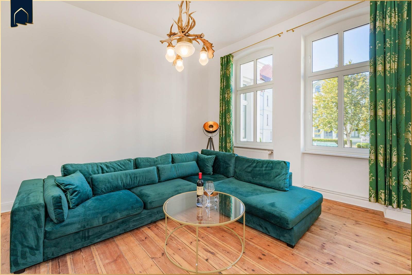 Ein Wohnzimmer mit grünem Sofa, Glas-Tisch und Fenstern mit grünen Vorhängen.