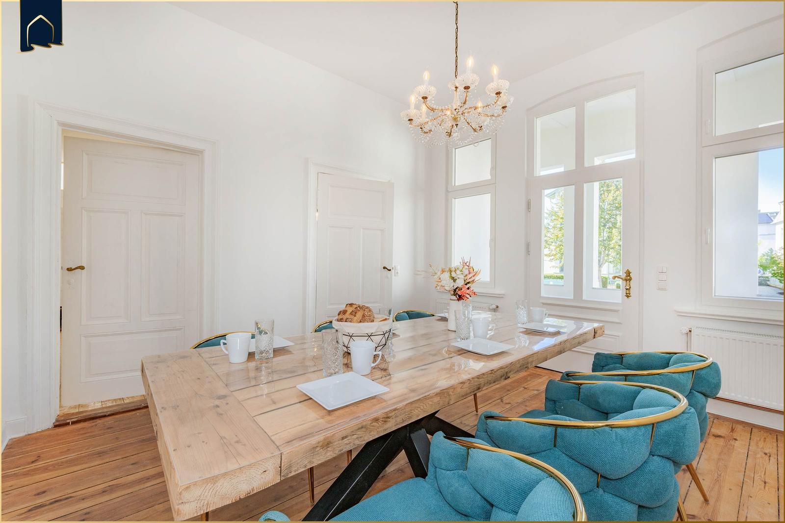 Esszimmer mit Holztisch, blauen Stühlen und Kronleuchter. Große Fenster und helle Wände.