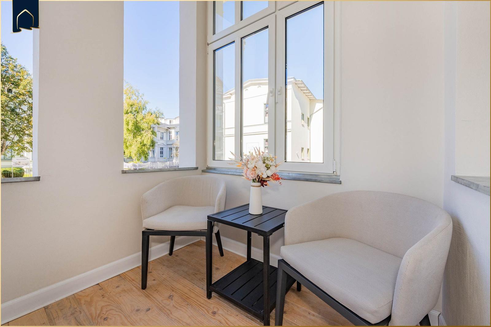 Zwei Sessel und ein Tisch stehen vor großen Fenstern mit Blick auf Gebäude.