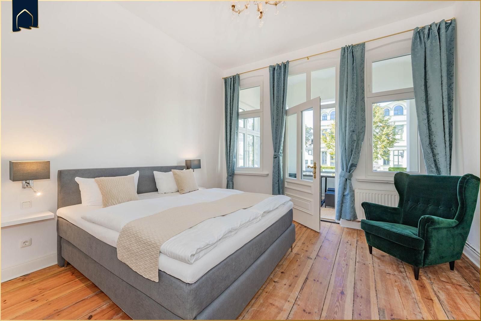 Ein Schlafzimmer mit Doppelbett, grünem Sessel und Holzboden. Fenster mit blauen Vorhängen.