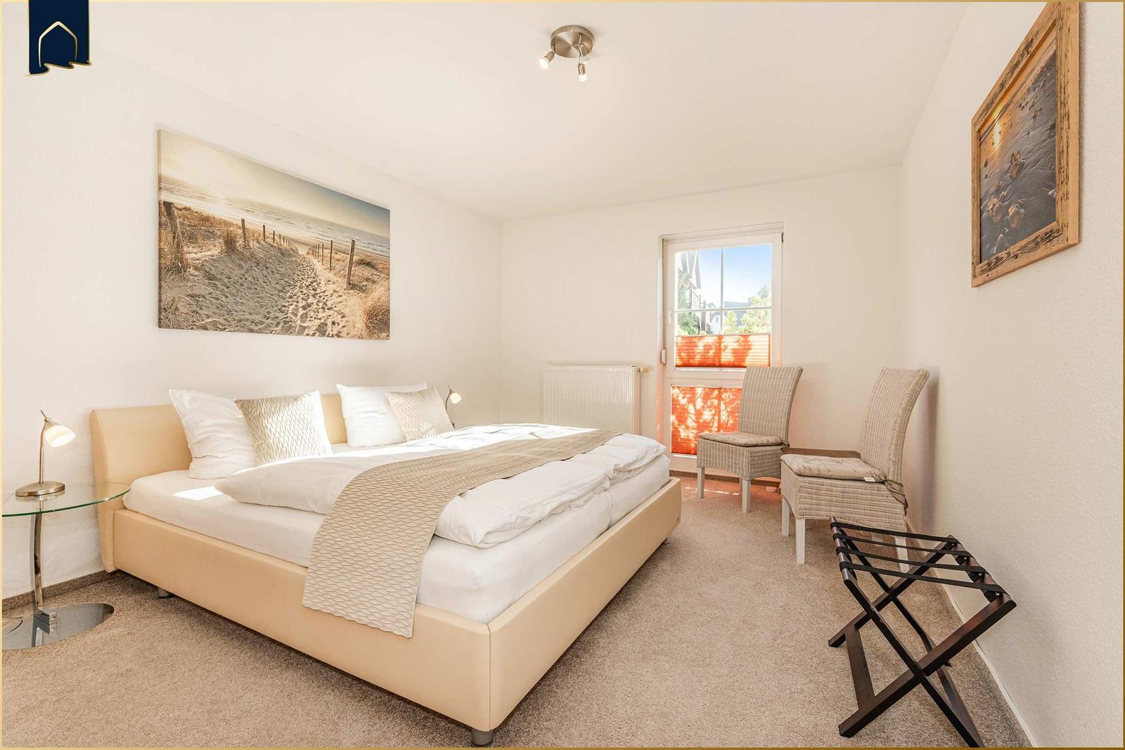 Schlafzimmer mit Bett, zwei Stühlen und Fenster. Wandbilder und Teppichboden.