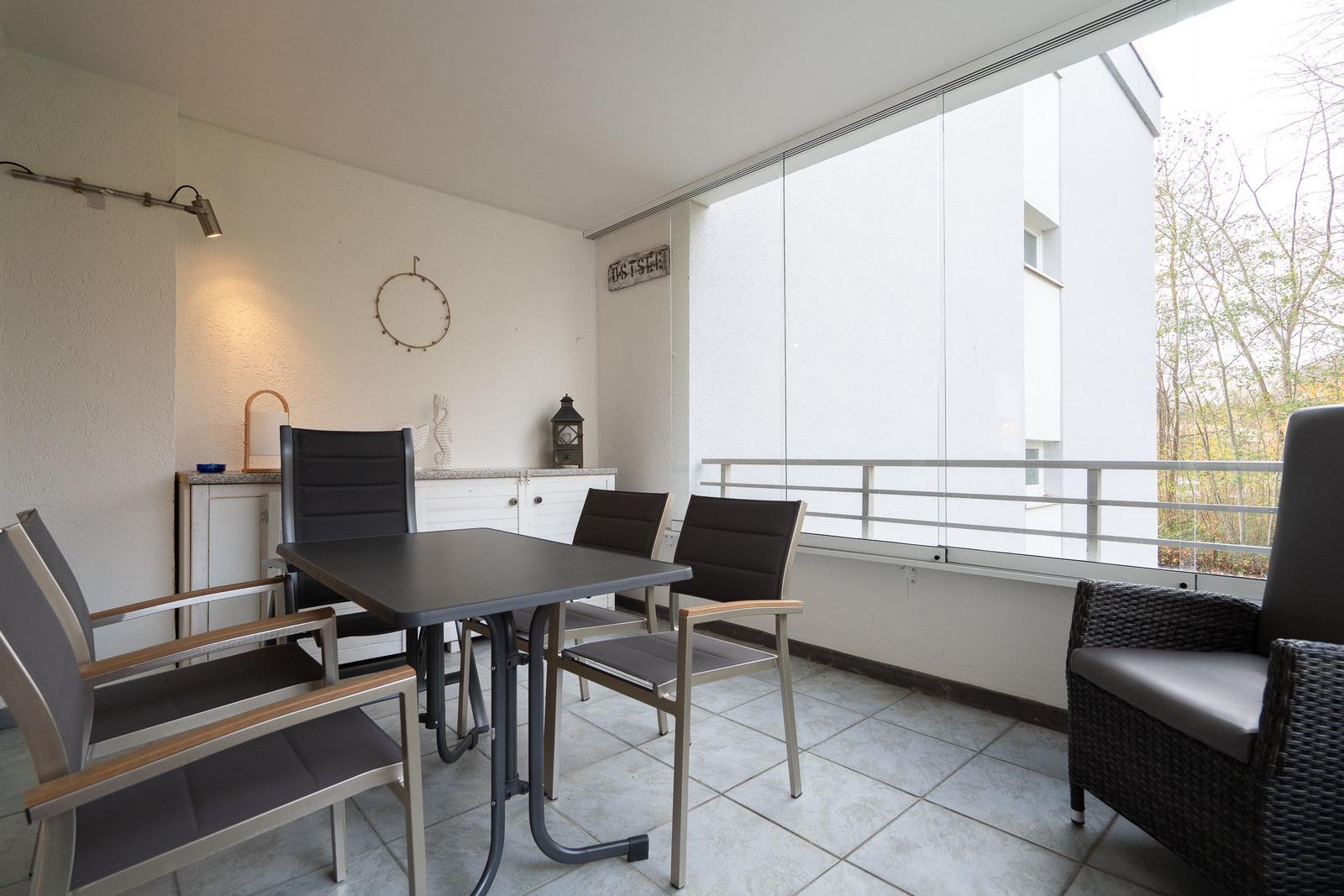 Terrasse mit Tisch, Stühlen und Sitzgelegenheit. Glaswand und Blick auf Bäume.