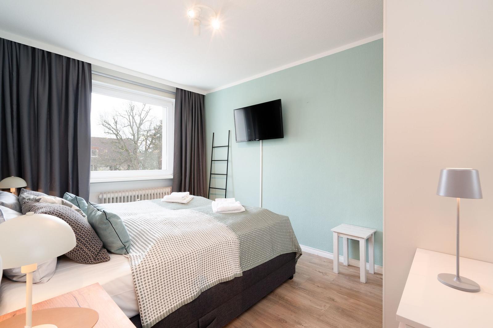 Schlafzimmer mit Bett, TV und Fenster. Holzboden und hellblaue Wand.