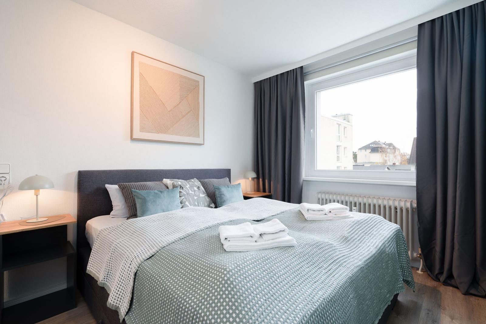 Schlafzimmer mit Bett, Nachttischen und Fenster mit Blick auf Gebäude.