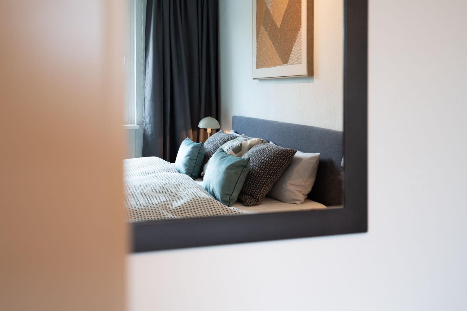 Ein Bett mit Kissen und Decke ist im Spiegel zu sehen. Dunkle Vorhänge und Wandbild sind sichtbar.