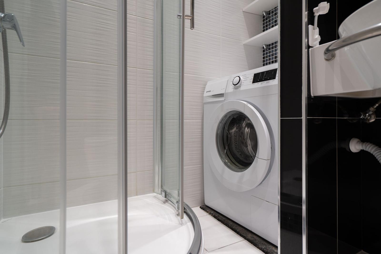 Dusche mit Glaswand, Waschmaschine und Waschbecken in moderner Badezimmerausstattung.