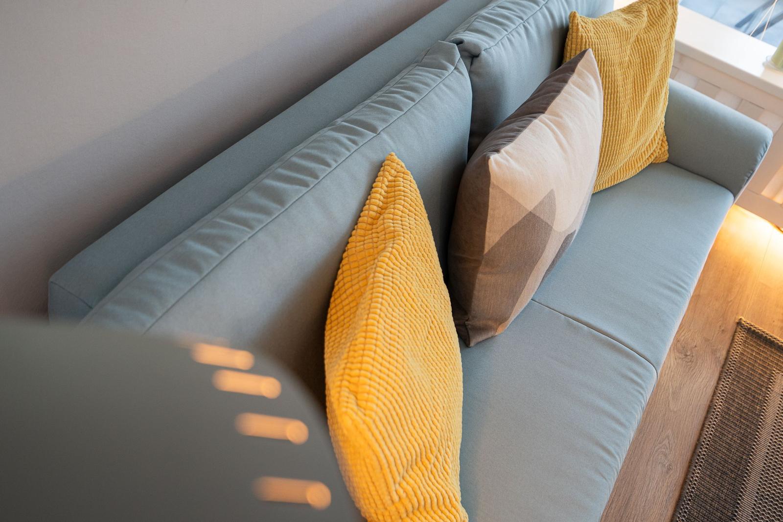 Ein hellblauer Sofa mit gelben und grauen Kissen. Holzboden und Fensterblick.
