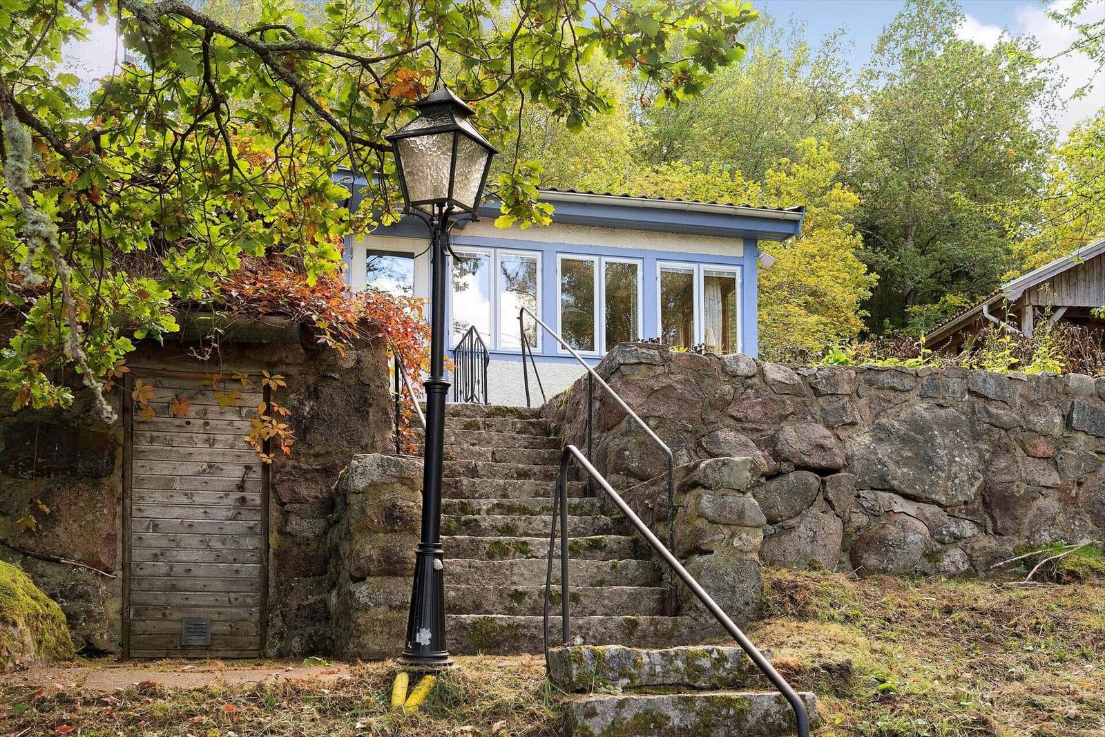 Ein blaues Haus mit Fenstern, Zugang über Steintreppe und Laterne. Umgeben von Bäumen und Steinmauer.