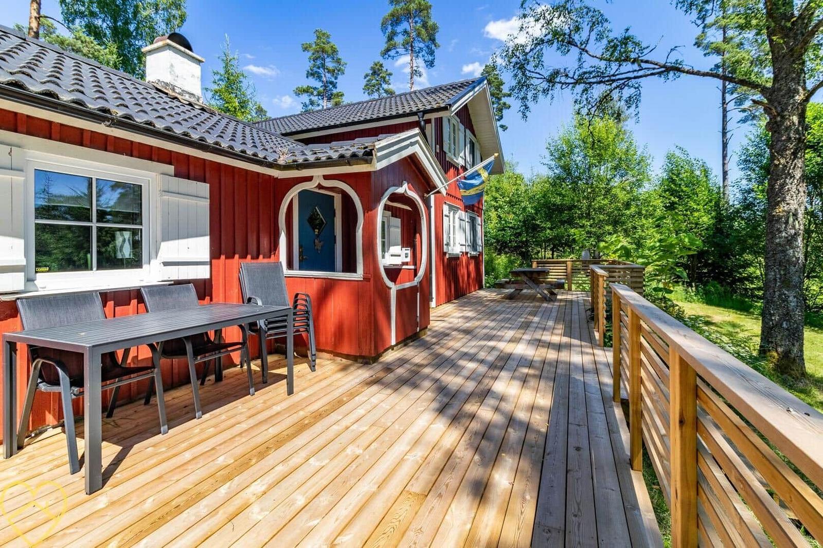 Rotes Haus mit Holzterrasse, Tisch und Stühlen, umgeben von Bäumen.