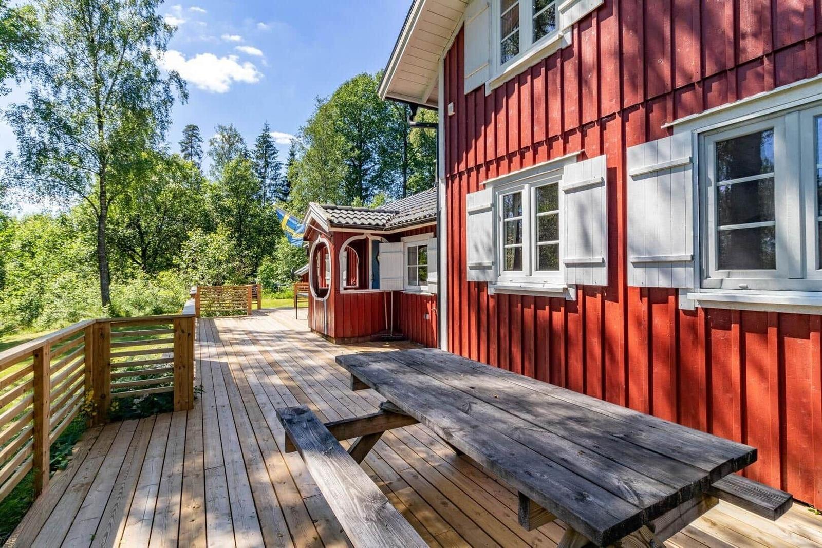 Holzterrasse mit Tisch vor rotem Haus, umgeben von Bäumen.