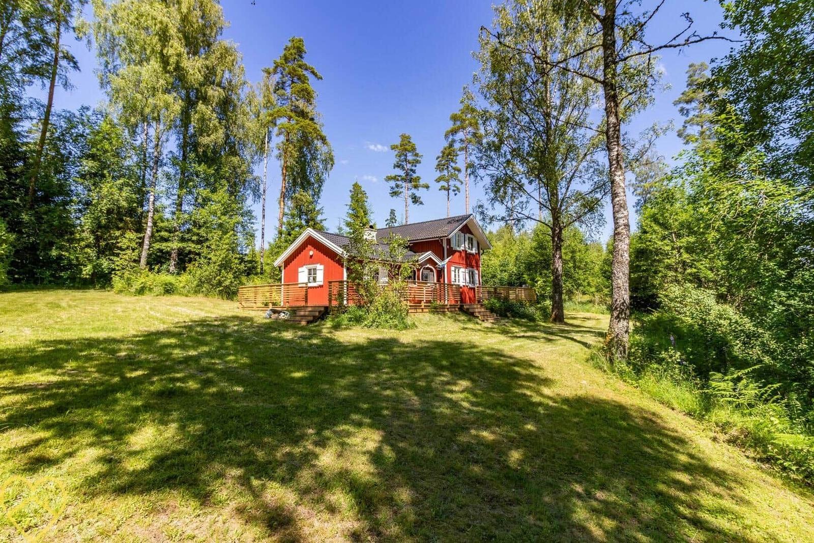 Rotes Holzhaus mit Terrasse im Wald. Grünflächen und hohe Bäume umgeben das Haus.