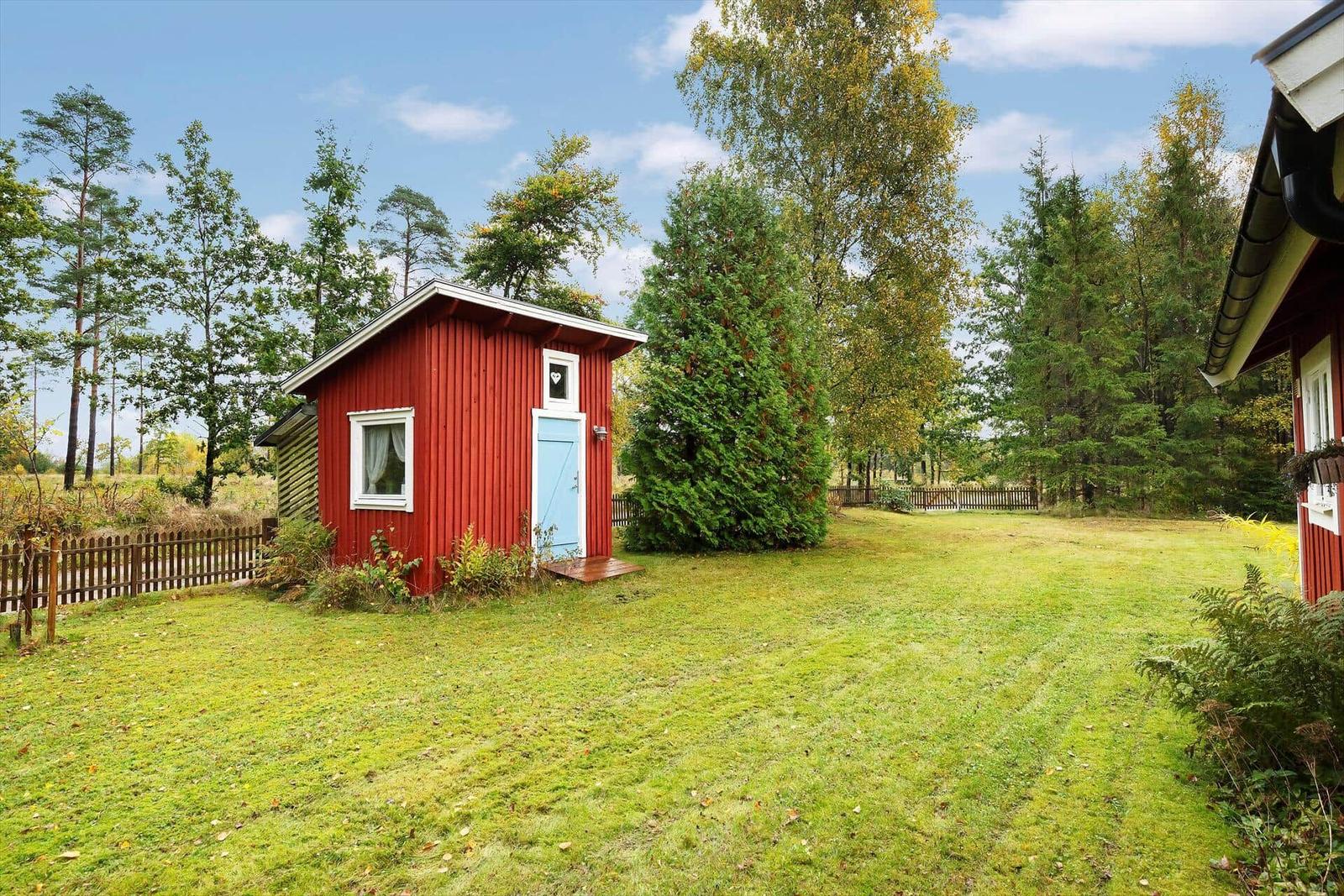 Rotes Holzhaus mit blauer Tür im grünen Garten. Umgeben von Bäumen und einem Zaun.