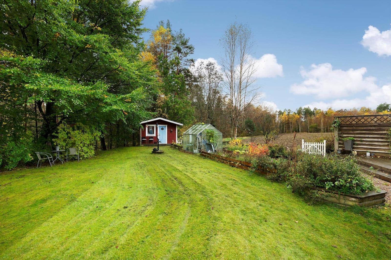 Roter Holzschuppen mit Garten, Gewächshaus und Terrasse unter blauem Himmel.