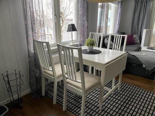 Esszimmer mit weißem Tisch und Stühlen, Fenster mit Vorhängen, Sofa im Hintergrund.