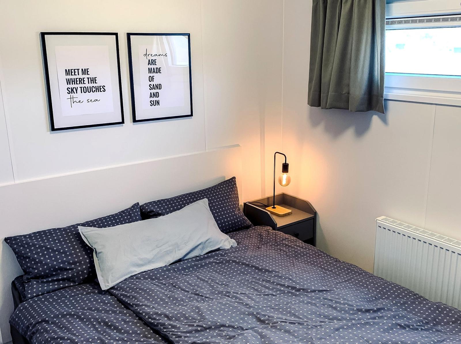 Schlafzimmer mit Bett, Nachttisch und Fenster. Wandbilder und Lampe sind sichtbar.