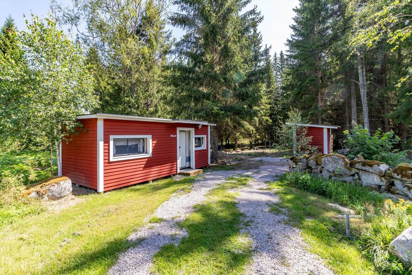 Rotes Holzhaus mit weißem Rahmen im Wald. Kiesweg führt zur Tür.