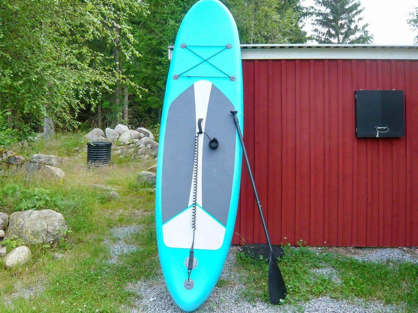 Ein türkisfarbenes Stand-Up-Paddleboard mit Paddel steht vor einem roten Schuppen.