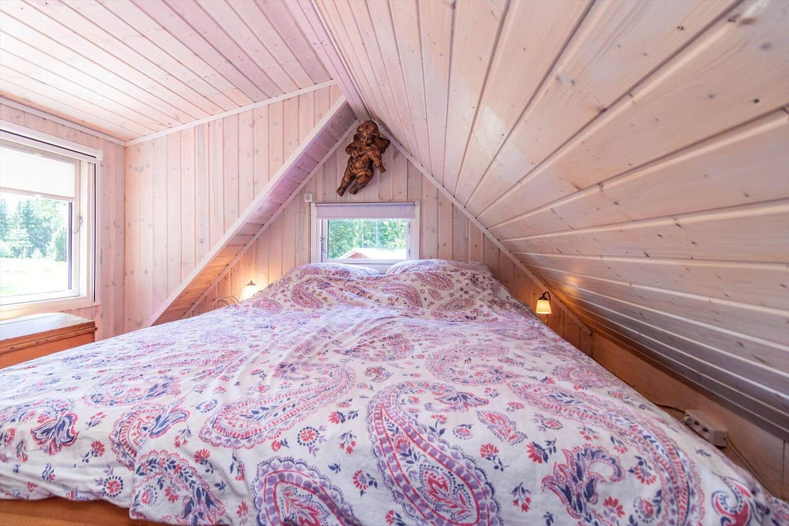 Schlafzimmer mit Holzwand und Dachschrägen, Bett mit Muster, Fenster und Wandlampe.