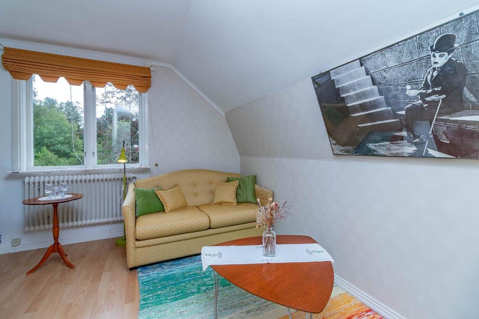 Wohnzimmer mit Sofa, Tisch und Wandbild. Fenster mit Vorhang. Holzboden und Teppich.