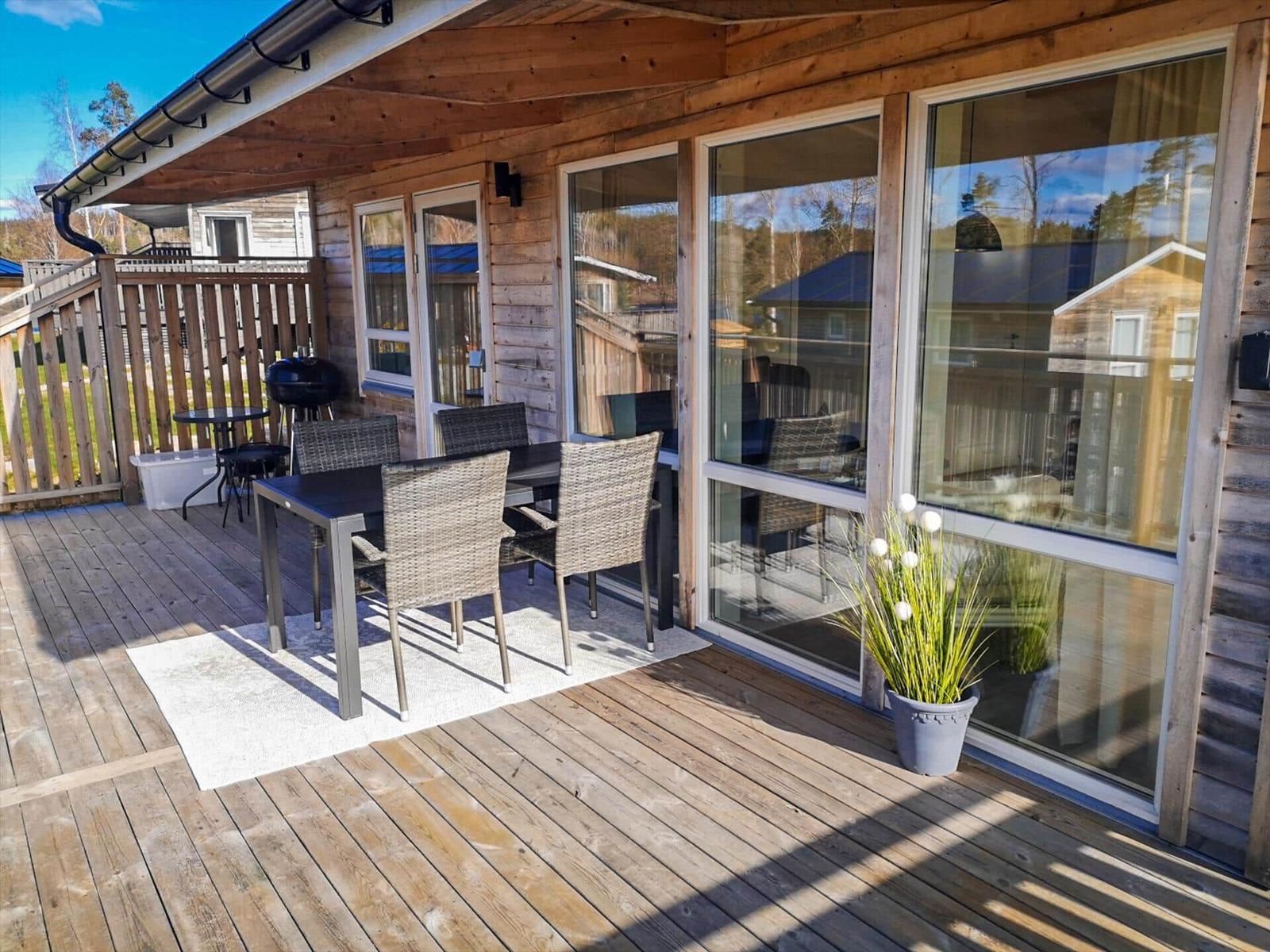 Terrasse mit Sitzgruppe, Grill und Pflanze vor Holzhaus mit Glasfront.