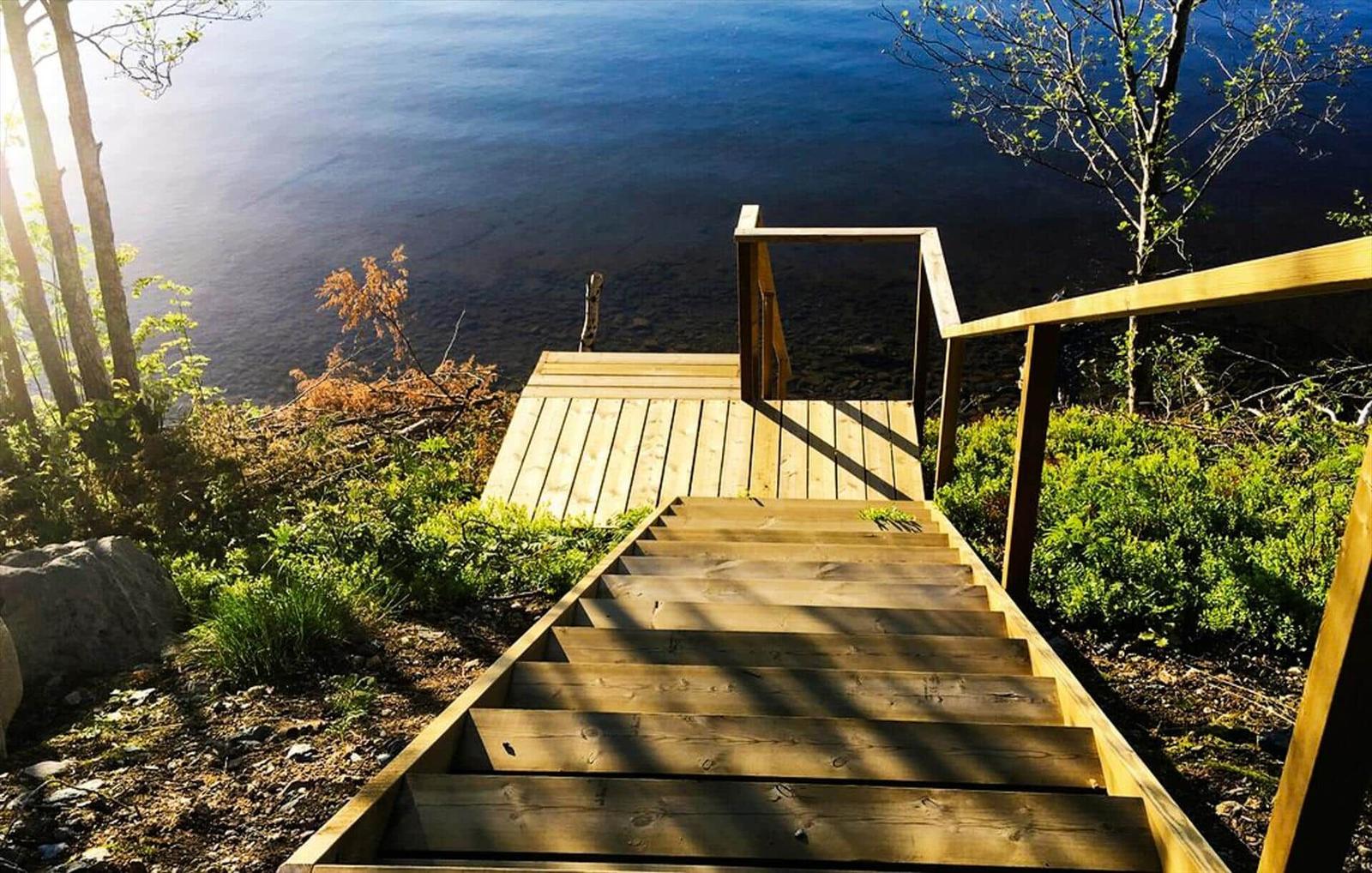 Holzstufen führen zu einer Terrasse am See. Umgeben von Bäumen und Grün.