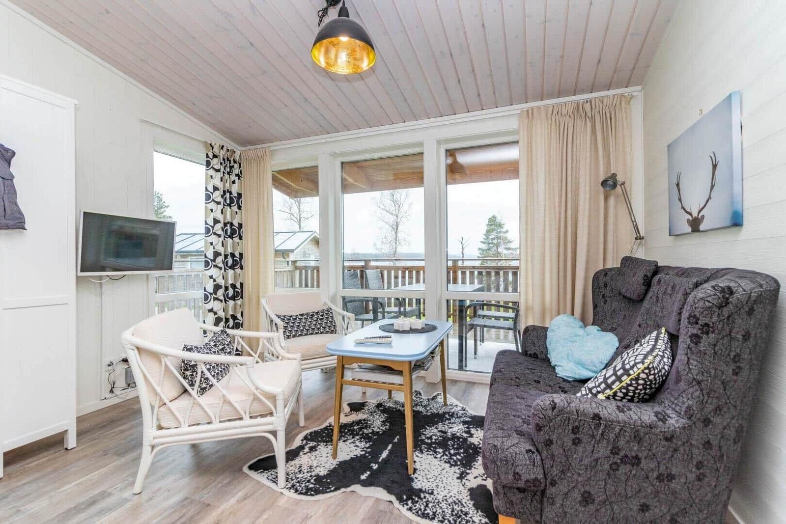 Wohnzimmer mit Sofa, Stühlen und TV. Balkon mit Sicht auf Wald.