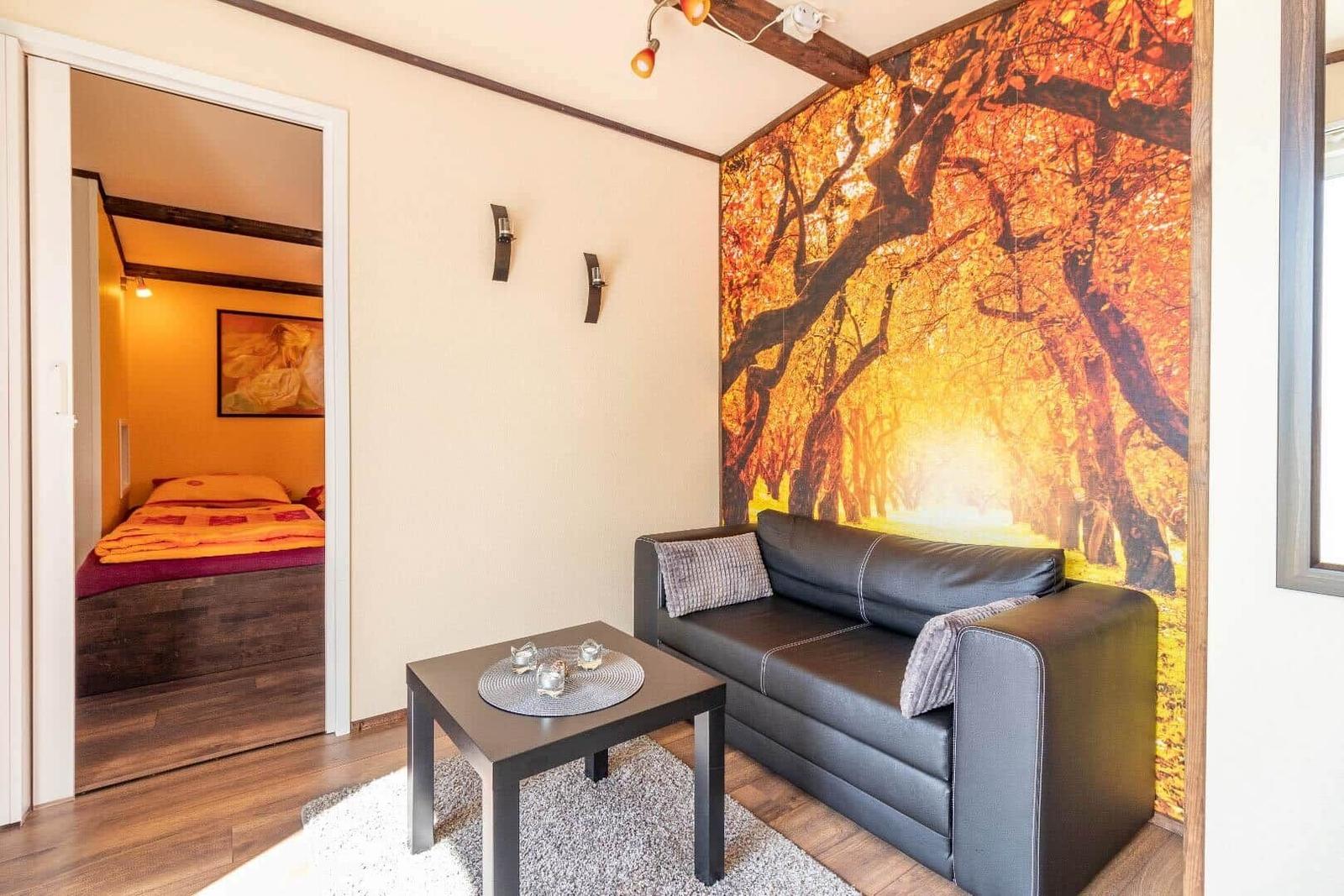 Wohnbereich mit Sofa und Tisch. Wandtapete mit Baummotiv. Tür zum Schlafzimmer.