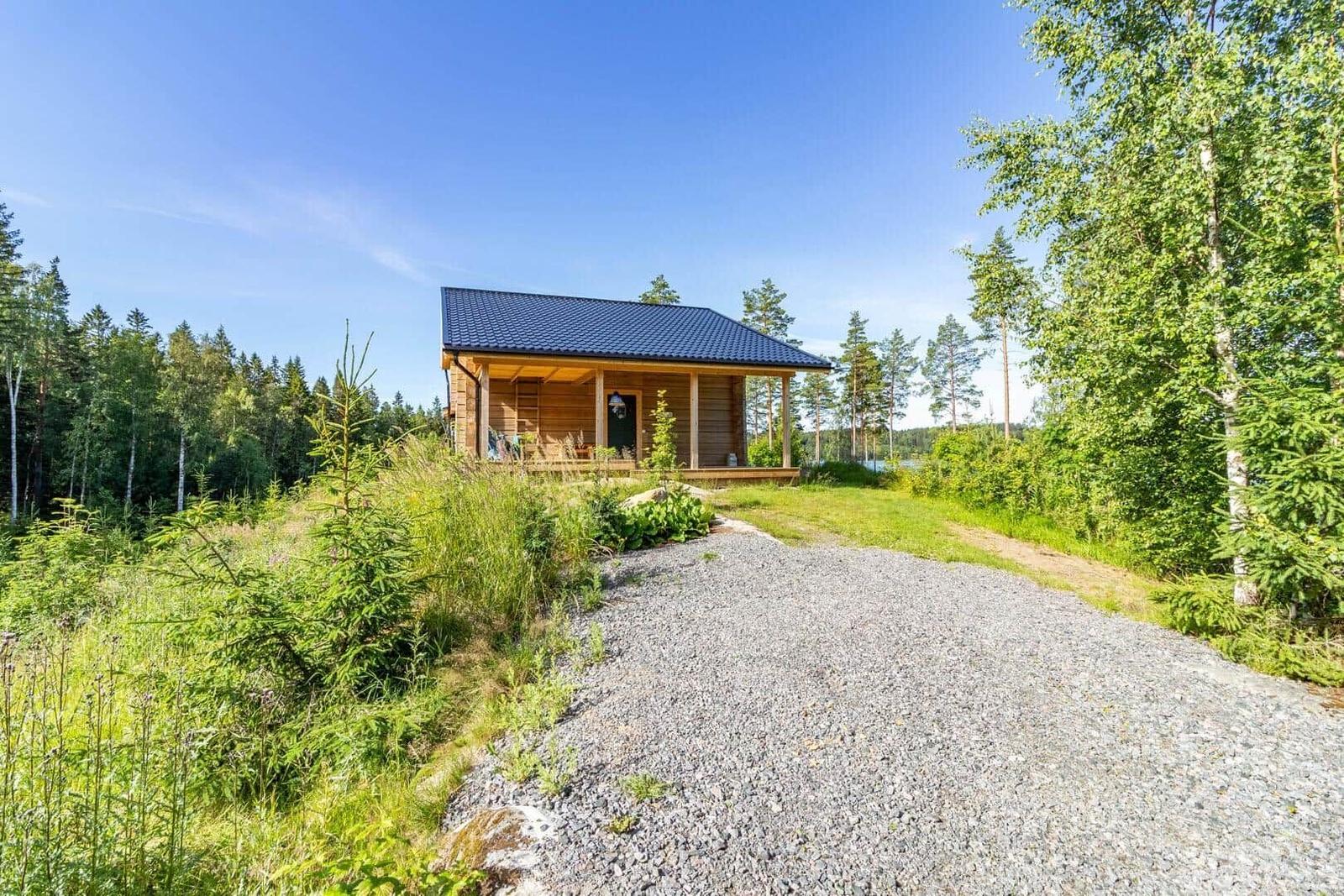 Wooden cabin with porch, surrounded by forest and grassland under blue sky.