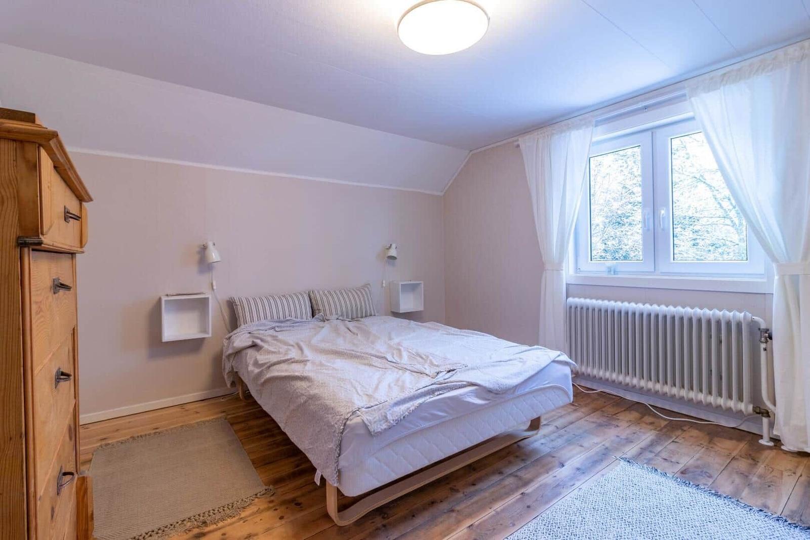 Schlafzimmer mit Bett, Holzboden und Fenster mit Blick auf Bäume.