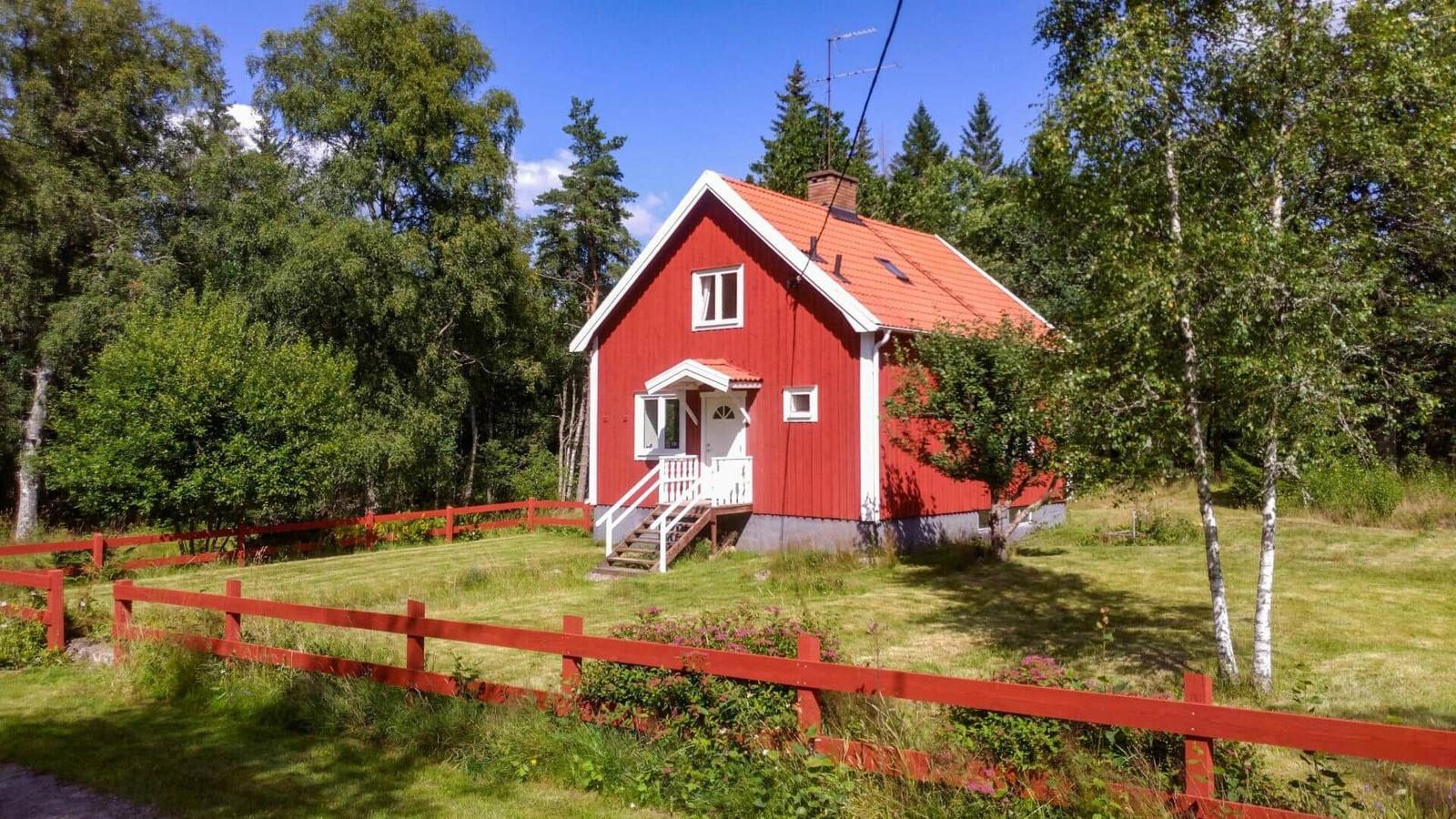 Rotes Holzhaus mit weißen Fensterrahmen und rotem Zaun im Grünen.