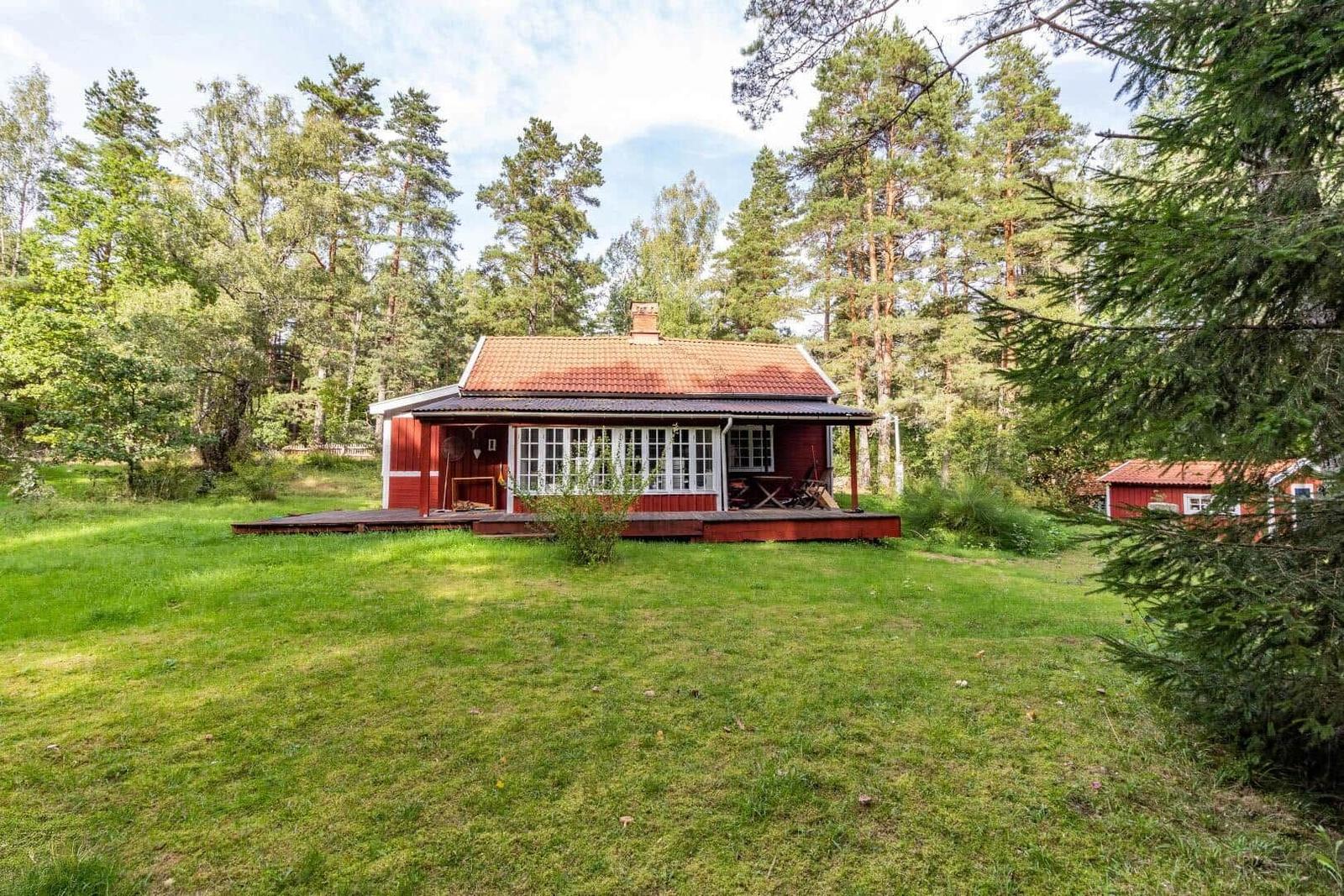 Red wooden house with terrace in the forest, green lawn and tall trees surround.