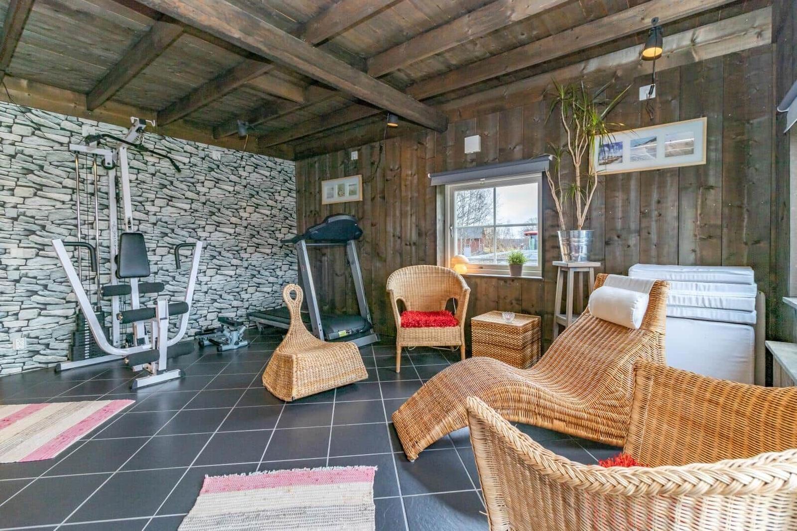 Fitnessraum mit Laufband, Kraftgerät und Rattanmöbeln. Holzwände und Steinwand.