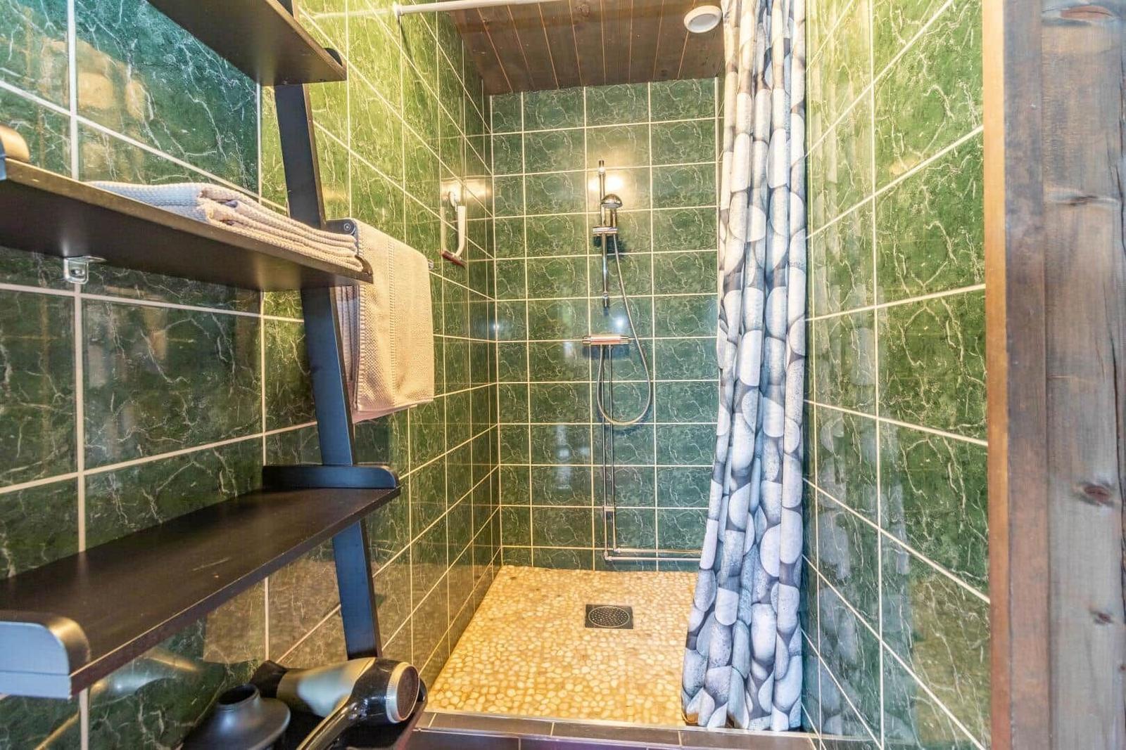 Dusche mit grünem Fliesenspiegel, Steinboden und Duschvorhang mit Steinmuster.